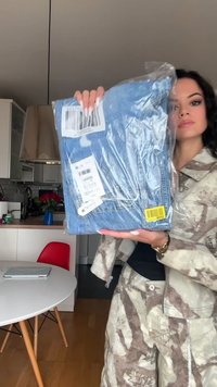 Frau in gemusterter Jacke und Hose hält verpackte blaue Jeans mit Barcode-Etiketten in einer modernen Küchenszene.
