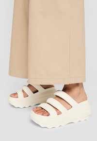 Weiße Plateau-Sandalen mit drei breiten Riemen, aus weichem Material. Die Sohle hat ein welliges Design, das einen strukturierten Halt bietet.