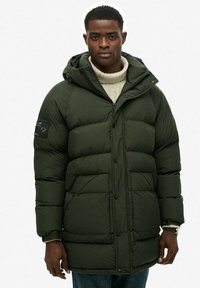 Chaqueta acolchada de color verde oscuro con cierre de cremallera y botón, con capucha y bolsillos laterales. La tela parece suave con un patrón acolchado.