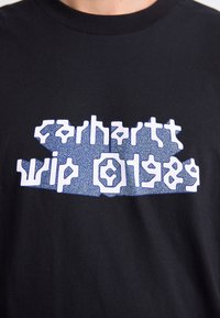 Person iført en sort skjorte med stiliseret hvid og blå tekst, der skriver "Carhartt WIP" på brystet.