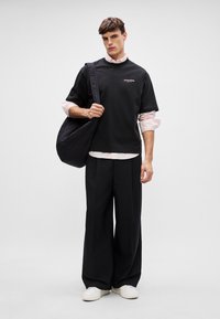 Camisa preta de manga curta com o colarinho redondo, combinada com calças largas pretas e ténis brancos. Uma grande mala preta está pendurada a um dos ombros.