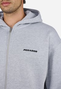 Szürke zipzáros kapucnis pulóver sima anyagból, elülső cipzárral és fekete "PEGADOR" logóval a bal mellkasi részen.