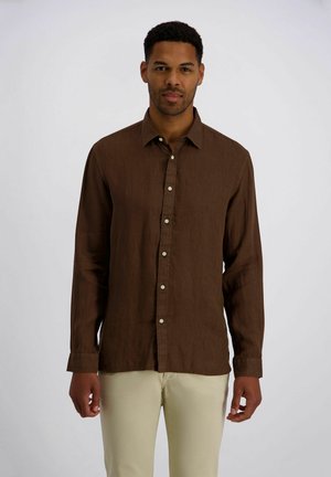 Mann trägt ein braunes, langärmliges Hemd mit Knöpfen und beige Hosen, steht vor einem schlichten hellgrauen Hintergrund.