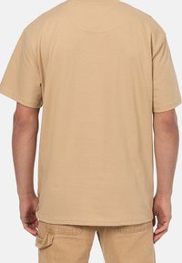 Krátký béžový tričko z bavlny s kulatým výstřihem a volným střihem, s viditelnými švy na zadní straně.