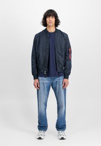 Marine bomberjakke med glidelås, ribbede mansjetter og sidelommer; brukt over en marineblå skjorte og paret med falmede blå jeans og sneakers.