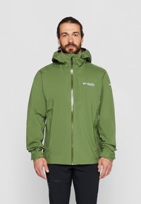 Hombre con barba que lleva una chaqueta verde Columbia Titanium con capucha, pantalones negros, de pie contra un fondo claro y liso.
