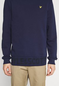 Sudadera de punto azul marino con mangas largas, puños y cintura acanalados, que presenta la inscripción en negro "LYLE & SCOTT" y el logotipo del pájaro amarillo.