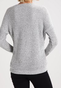 Femme portant un pull à manches longues, gris clair, en maille avec un motif texturé, associée à un pantalon noir, vue de dos.