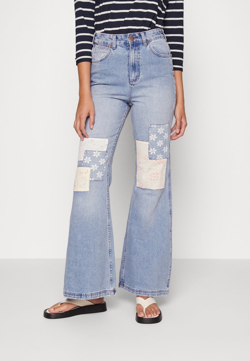 Personne portant un jean évasé taille haute bleu clair avec des motifs floraux pastel et des sandales noires et blanches, associé à un haut à rayures noires et blanches.