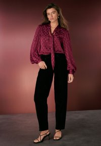 Blusa color bordeaux con motivo a pois e collo con fiocco, abbinata a pantaloni in velluto neri a vita alta e sandali neri con cinturini. Texture lisce.