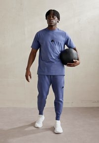 Niebieska koszulka sportowa z krótkim rękawem i czarnym logo, dopasowane dresy oraz białe sneakersy. Trzyma czarną piłkę do koszykówki.