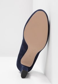 Escarpin en daim bleu marine avec un bout pointu et un talon fin. La semelle est en un matériau léger et lisse, avec un logo de la marque gravé.