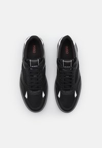 Baskets noires avec une tige en maille et synthétique, bout rond, détails texturés, accents blancs et lacets noirs. Logo de la marque sur la languette.