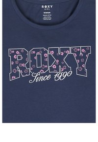 Navy organic cotton T-shirt featuring "ROXY" in white outline filled with pink floral pattern and "Since 1990" in white cursive text.