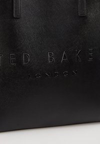 Czarna torba na ramię z teksturowanego materiału syntetycznego, z wytłoczonym napisem "TED BAKER" i "LONDYN". Solidne uchwyty, prostokątny kształt.