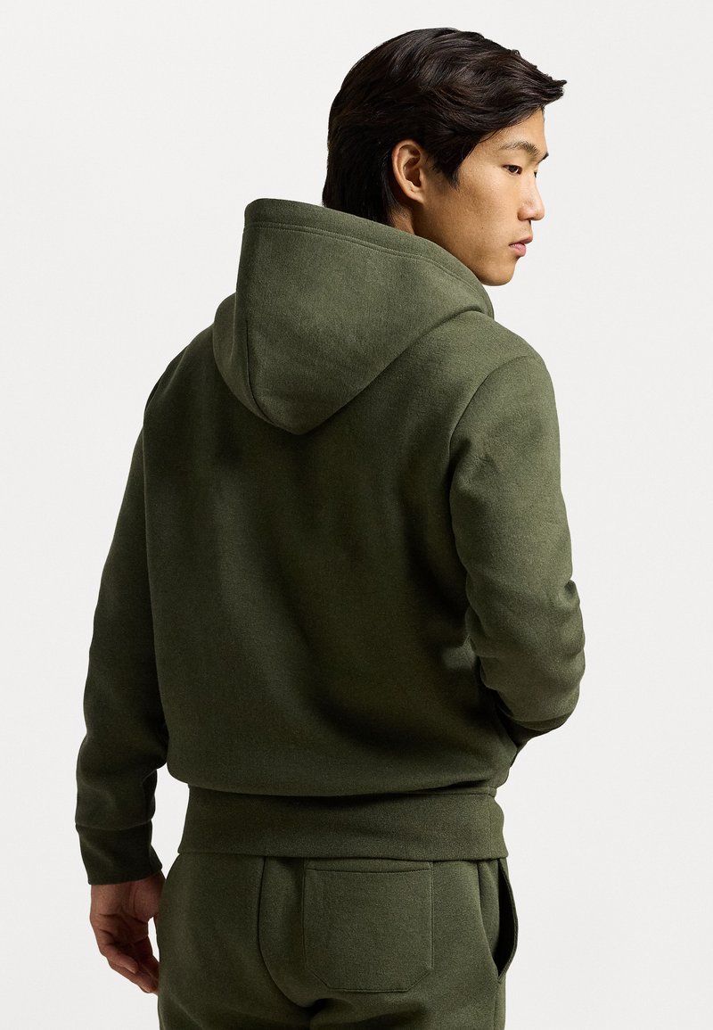 Sudadera verde oliva hecha de una tela de algodón suave y gruesa, con una capucha espaciosa y puños acanalados, diseñada con un ajuste holgado.