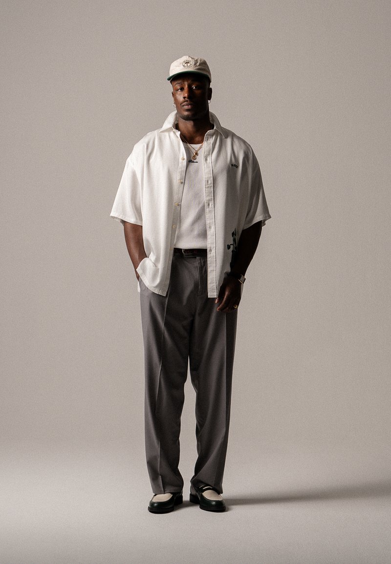 Chemise blanche à manches courtes sur un débardeur blanc, pantalon gris et chaussures noires avec des accents blancs. Accessoire comprenant une casquette et des bijoux.