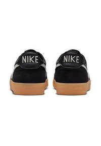 Las zapatillas Nike de ante y cuero negro presentan una suela de goma color caramelo, con la marca "NIKE" en el talón y una franja en blanco contrastante en la entresuela.