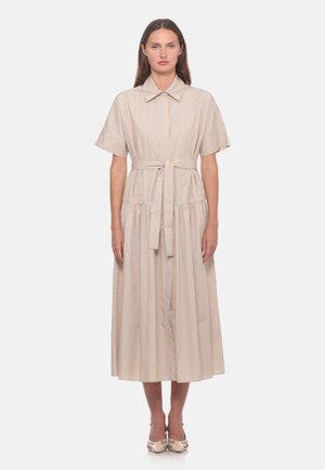 Donna che indossa un vestito midi beige a maniche corte con colletto, sottili righe verticali e cintura in vita, in piedi dritta su uno sfondo bianco.