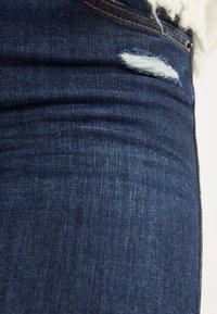 Primer plano de unos jeans de mezclilla azul oscuro con un pequeño desgarro deshilachado cerca de la cinturilla y costuras naranjas visibles.