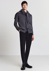 Maglione grigio scuro con collo sciallato, chiusura con bottoni e due tasche frontali, abbinato a pantaloni neri slim-fit e stivali neri lucidi.