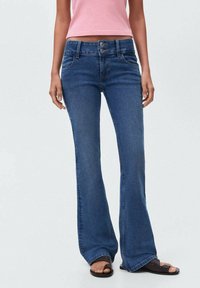 Jeans de mezclilla acampanados en azul oscuro con cierre de dos botones, cinco bolsillos y una textura suave. Combinados con sandalias negras.