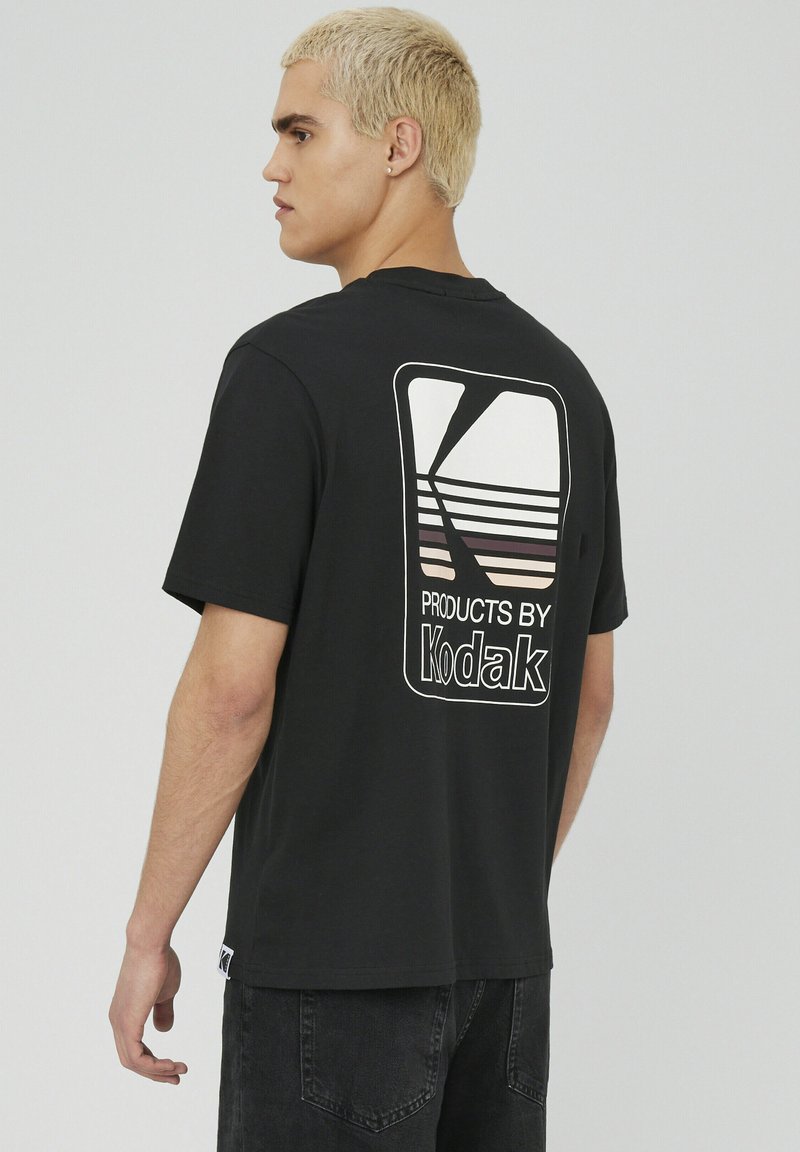 T-shirt en coton noir avec un grand logo Kodak blanc sur le devant, et des lignes horizontales dans le dos. Manches courtes et coupe décontractée.