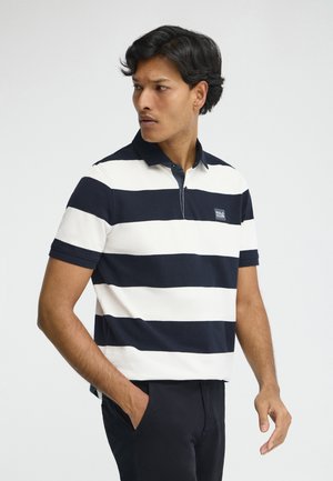 Hombre que lleva una camisa polo de rayas anchas en blanco y negro, con mangas cortas, de pie con una mano en el bolsillo, mirando hacia un lado.