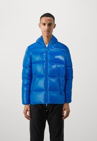 Lys blå pufferjakke med en blank finish, lynlåslukning foran og hætte. Har to sidelommer og en puflet segmenteret design.