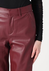 Pantalons en simili cuir bordeaux avec une texture lisse, dotés d'une taille ajustée, de poches latérales et de détails de couture visibles.