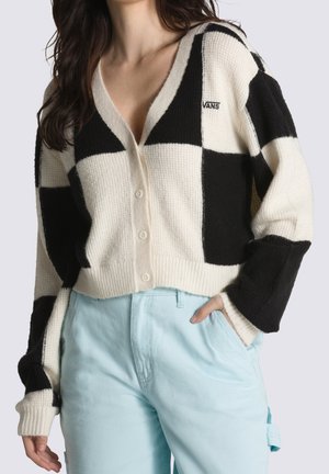 Gebreide cardigan met een zwart-wit ruitpatroon, v-hals, knoopsluiting en geribbelde boorden, gecombineerd met lichtblauwe broek.