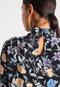 Femme aux cheveux bruns en queue de cheval, portant un blouse noire à fleurs avec un col volanté et un dos à bouton en trou de serrure, boucle d'oreille géométrique en or.