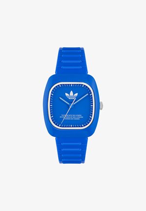 Čtvercové modré silikonové hodinky se shodným páskem. Mají plnou modrou ciferník, bílé hodinové značky a stříbrnou korunku s minimálním brandingem.