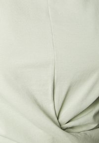 Světle zelený top vyrobený z vroubkované látky, který má na přední straně uzlový detail a hladký povrch s jemným leskem.