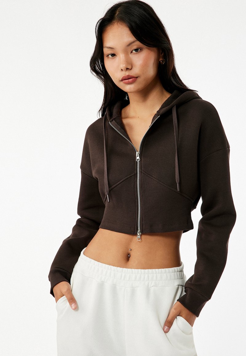Korte zip-up hoodie in donkerbruin, met een gestructureerde ribbelrand, voorzakken en een capuchon, gecombineerd met lichtgekleurde joggingbroek.