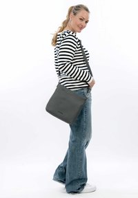 Borsa a tracolla in pelle grigia con una texture liscia, forma rettangolare e tracolla regolabile. Il modello indossa una camicia a righe bianche e nere e jeans.