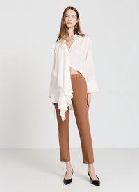 Weißes, rüschiges Oberteil mit Knopfschluss und braune, maßgeschneiderte Hose. Schwarze spitze High Heels runden das Outfit ab. Schlicher Hintergrund.