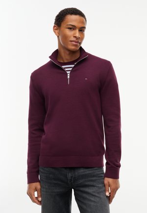 Joven con un jersey morado con cremallera hasta la mitad, sobre una camisa de rayas y vaqueros oscuros, apoyado sobre un fondo blanco liso.