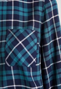 Tissu en tartan bleu et vert d'eau avec des lignes blanches, comportant une poche poitrine. Le tissu a une texture lisse, créant un design cohérent.