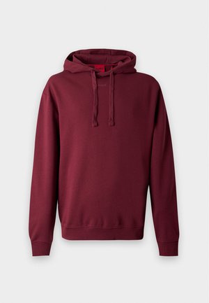 Homme portant un sweat à capuche bordeaux, un pantalon gris et des chaussures noires, debout avec une main dans la poche devant un fond uni.