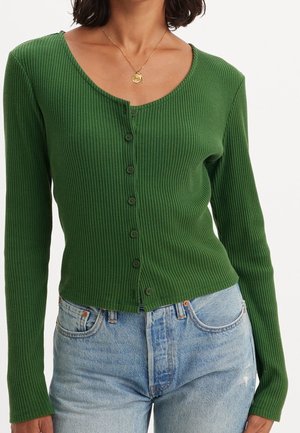 Femme portant un cardigan côtelé vert ajusté boutonné et un jean taille haute bleu clair, avec un collier pendentif en or.