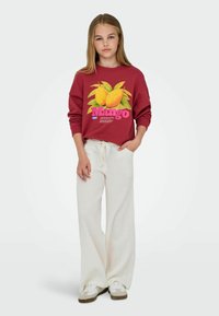 Bordo dukserica s grafikom manga i ružičastim tekstom, uparena s bijelim hlačama širokog kroja. Bijeli tenisice s sivoj akcentima upotpunjuju odjevnu kombinaciju.