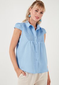 Blusa azzurro chiaro a maniche corte con chiusura a bottoni e dettagli in pizzo sul petto. Tessuto di cotone morbido con vestibilità comoda.