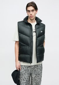 Mustast polsterdatud vest kõrge kaelusega, lukuga ja küljetaskutega. Kantud helevärvilise t-särgi peale ning kombineeritud mustriliste pükstega.