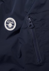 Navy windbreaker with a glossy texture, featuring a circular Kaporal patch and a visible zippered pocket. Zipper has a branded pull tab.