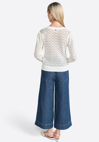 Maglione bianco lavorato a maglia con una trama aperta e testurizzata; polsini e orlo a coste. Abbinato a pantaloni in denim blu a gamba larga e scarpe di colore chiaro.