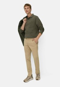 Olive green long-sleeve polo with pocket, beige trousers, and light brown sneakers. Model holds a dark green jacket over his shoulder.