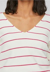 Sweter dzianinowy z dekoltem w kształcie litery V w kolorze białym z poziomymi różowymi paskami. Charakteryzuje się prążkowaną fakturą i luźnym krojem, podkreślając casualowy design.