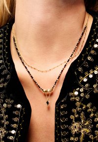 Colliers superposés en or et noir, ornés de délicates perles et d'un pendentif central en forme de goutte. Porté sur un cou nu.