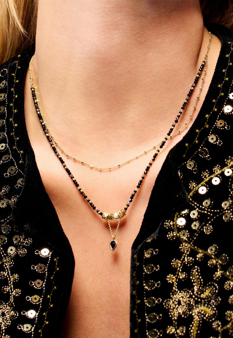 Colliers superposés en or et noir, ornés de délicates perles et d'un pendentif central en forme de goutte. Porté sur un cou nu.